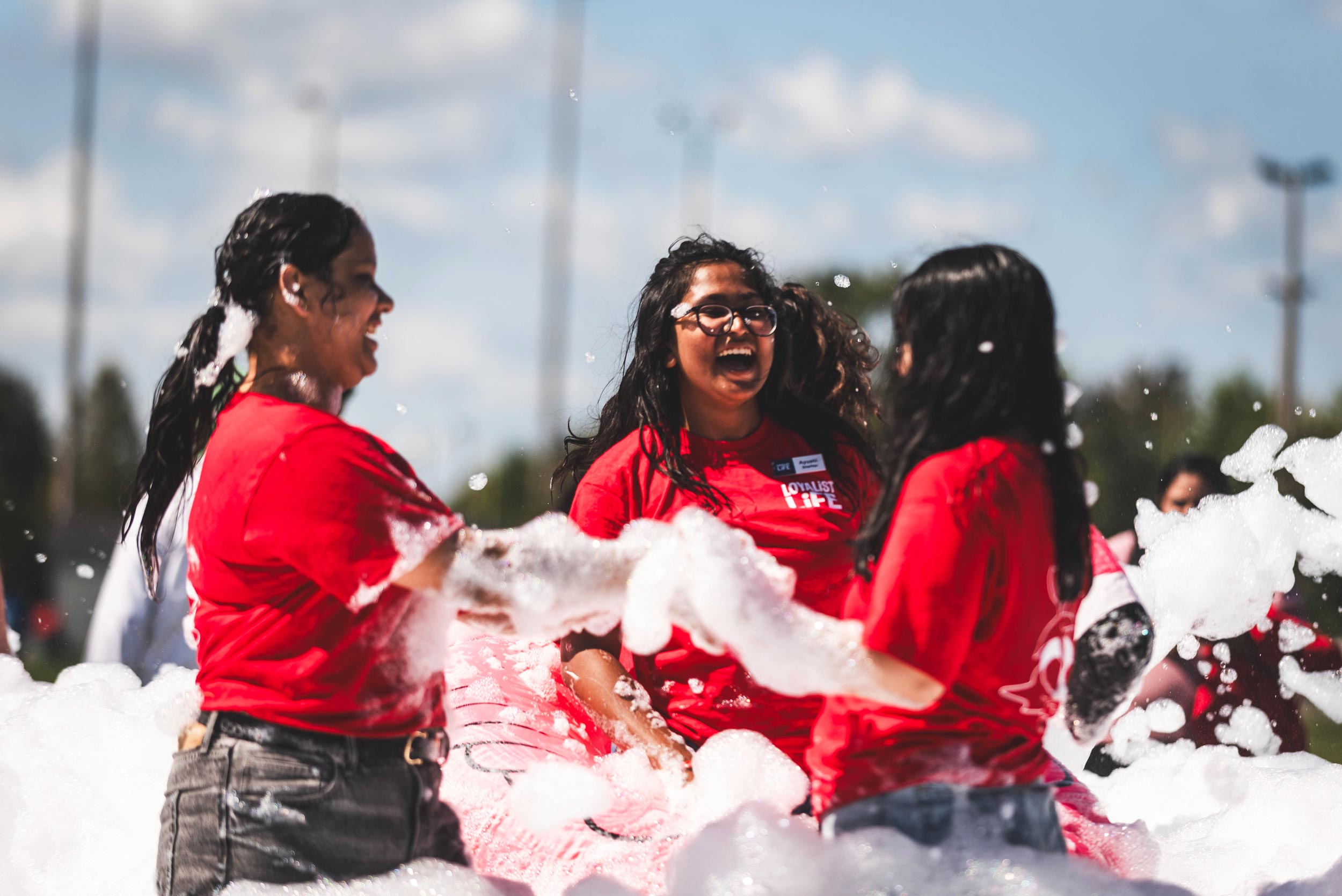 Three students wearing red Loyalist College shirts laughing and covered in foam during an outdoor event, enjoying the sunny day.