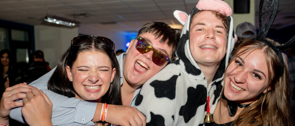 A group of four friends at a Halloween-themed pub night, smiling widely and wearing costumes. One person is dressed in a cow costume, while the others are in casual attire with playful accessories. They are embracing and clearly enjoying the festive atmosphere.