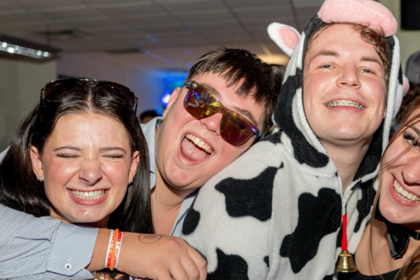 A group of four friends at a Halloween-themed pub night, smiling widely and wearing costumes. One person is dressed in a cow costume, while the others are in casual attire with playful accessories. They are embracing and clearly enjoying the festive atmosphere.