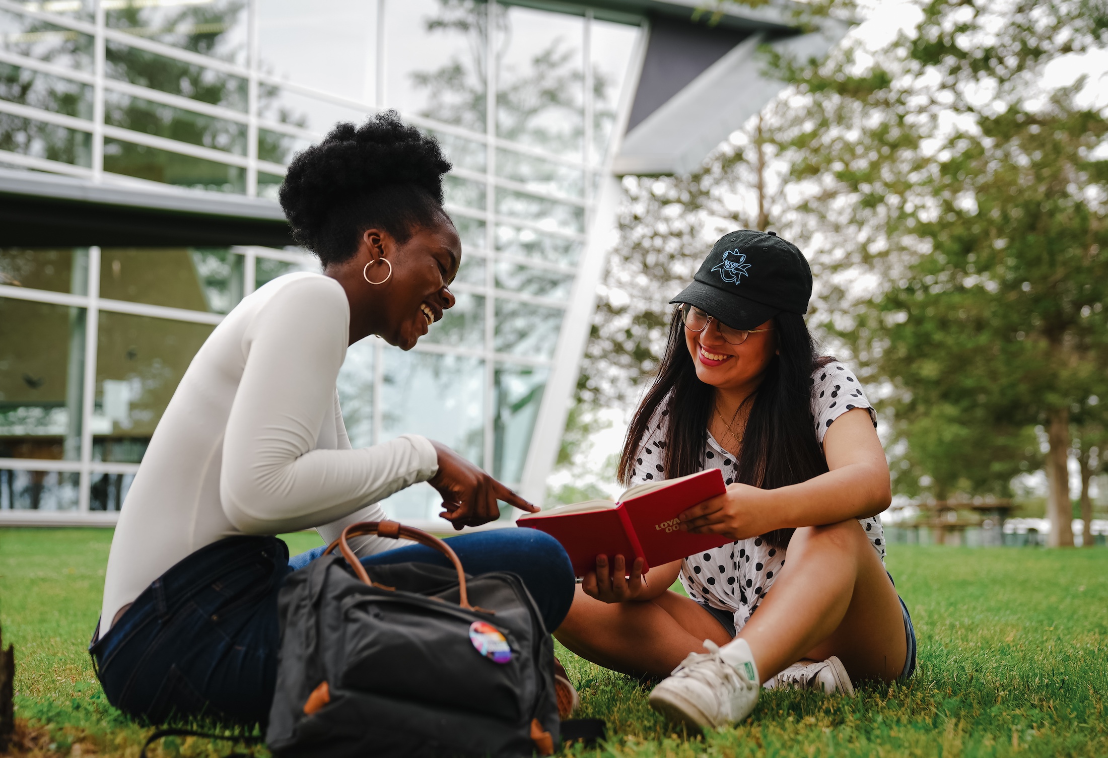 Two women sit on a grassy lawn in front of a modern building, engaging with a book. One points at the book while the other looks on, smiling. A backpack lies next to them.