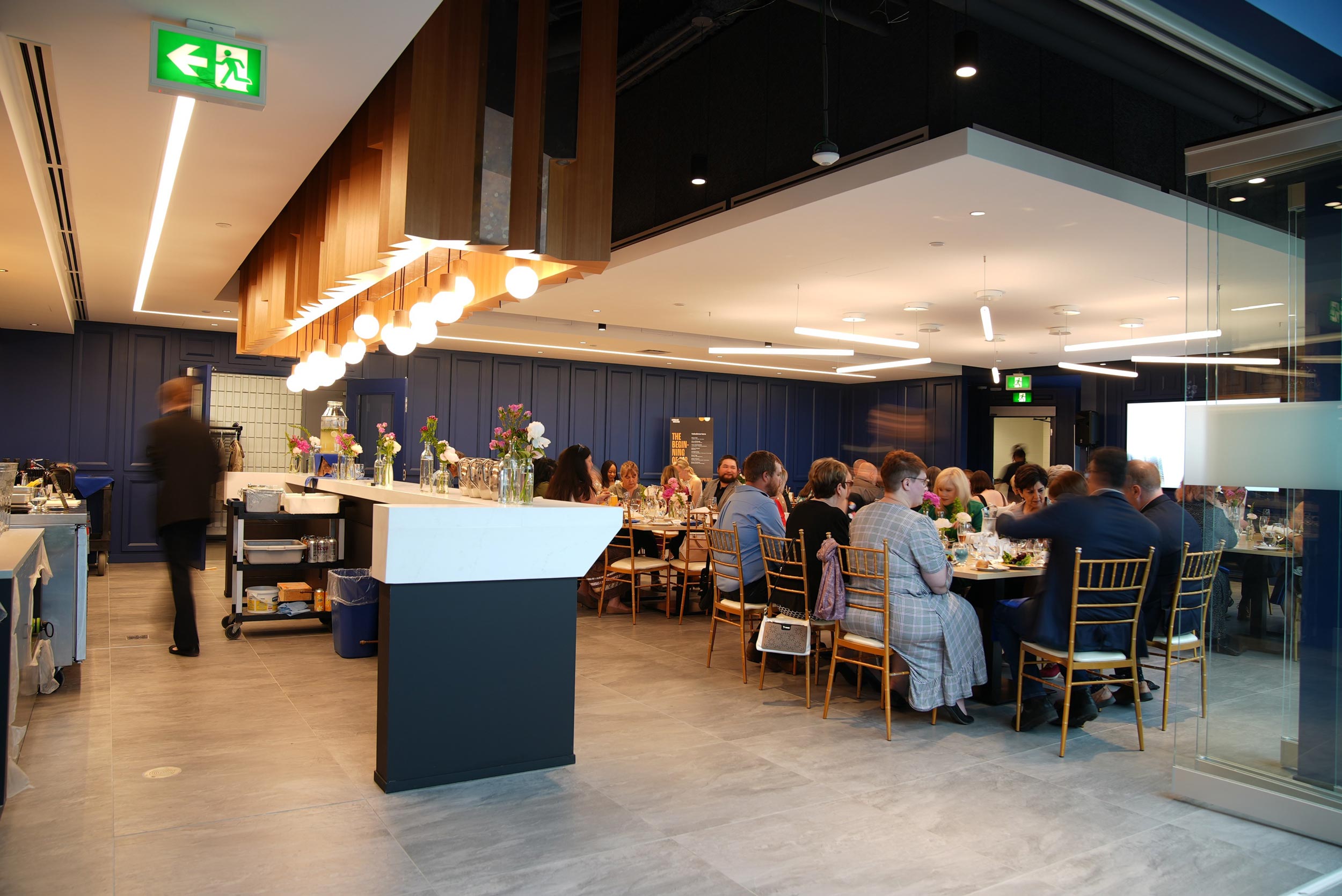 The interior of Loyalist College's Nourish restaurant. Guests sit at tables in the well-lit space and a waiter is in motion on the left side of the photo.