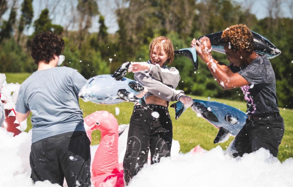 Three students playing in a foam party, smiling and playfully hitting each other with inflatable fish toys on a sunny day.