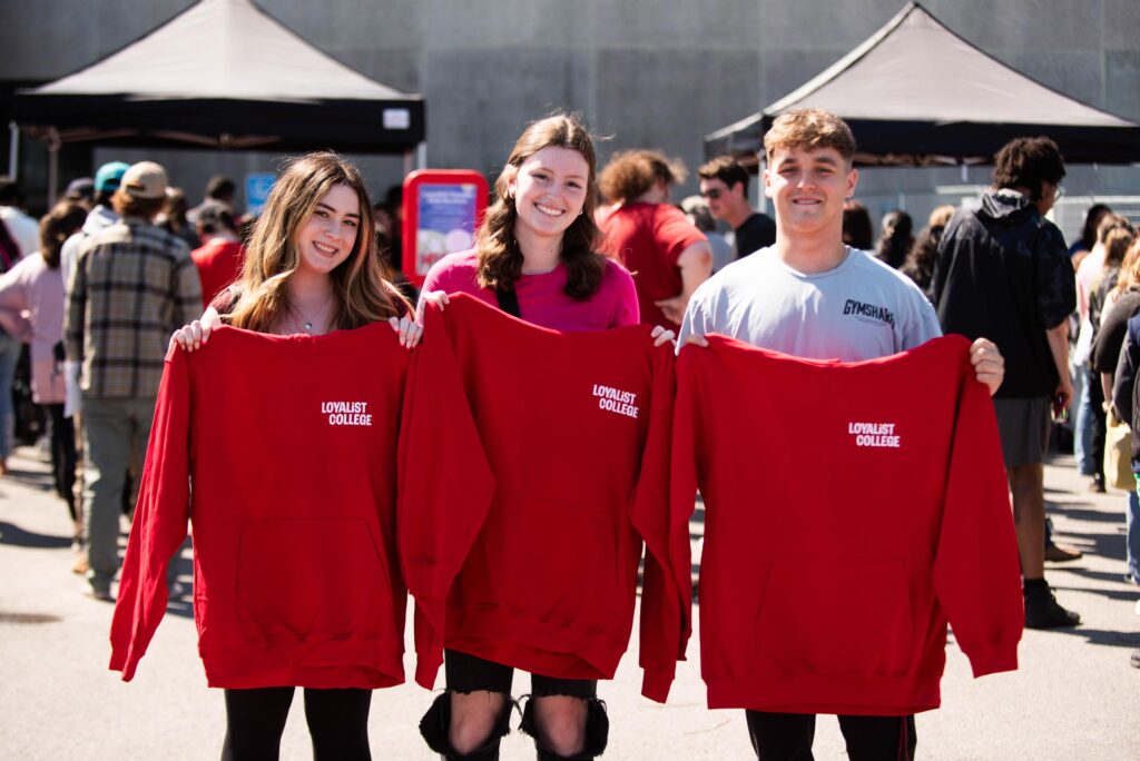 Three students smiling and holding up red Loyalist College sweatshirts during an outdoor event, with a crowd in the background.