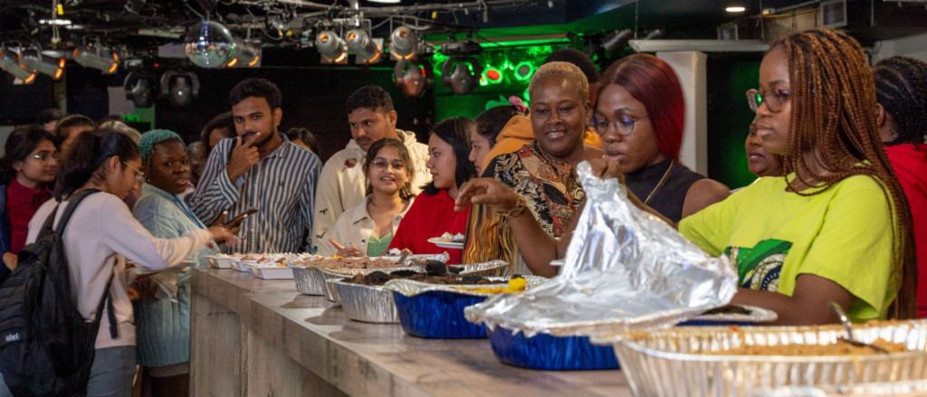 A diverse group of students standing in line at a buffet-style event. Various dishes are laid out in aluminum trays, and attendees are serving themselves food while engaged in conversation. The setting is a casual, well-lit room with vibrant green lighting and disco balls overhead, suggesting a celebratory or cultural event. The atmosphere is lively, with people smiling and enjoying the gathering.