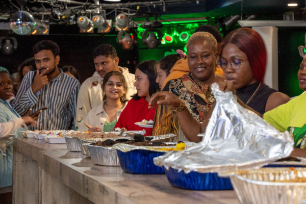 A diverse group of students standing in line at a buffet-style event. Various dishes are laid out in aluminum trays, and attendees are serving themselves food while engaged in conversation. The setting is a casual, well-lit room with vibrant green lighting and disco balls overhead, suggesting a celebratory or cultural event. The atmosphere is lively, with people smiling and enjoying the gathering.