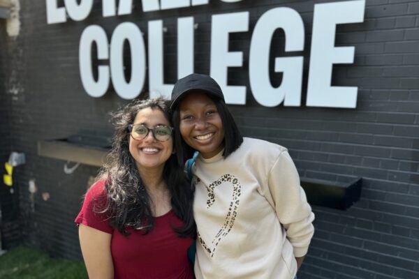 Two students smiling and posed in front of the Loyalist College logo on the Port Hope campus.