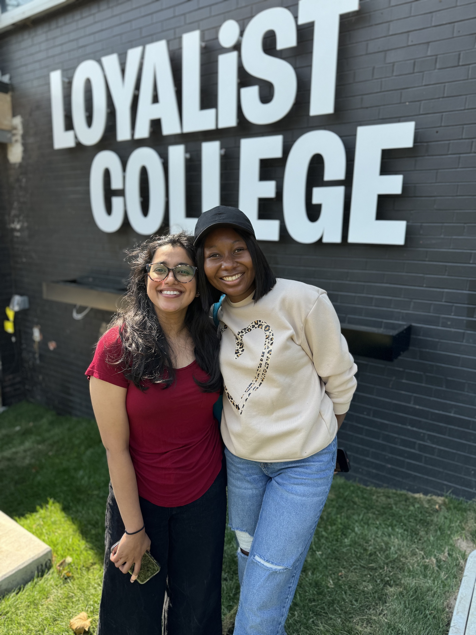 Two students smiling and posed in front of the Loyalist College logo on the Port Hope campus.