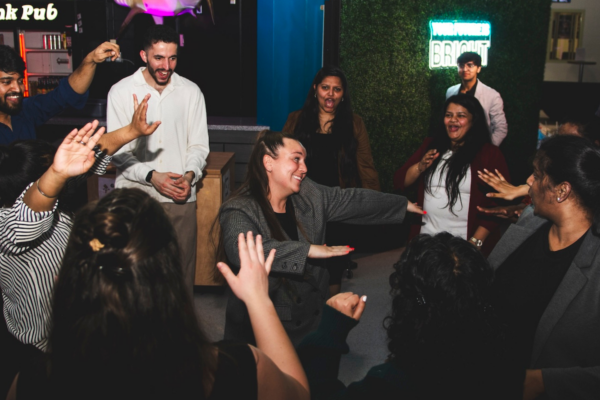 A lively group of people dancing and laughing inside a pub. A neon sign reading "Shark Tank Pub" glows in the background.