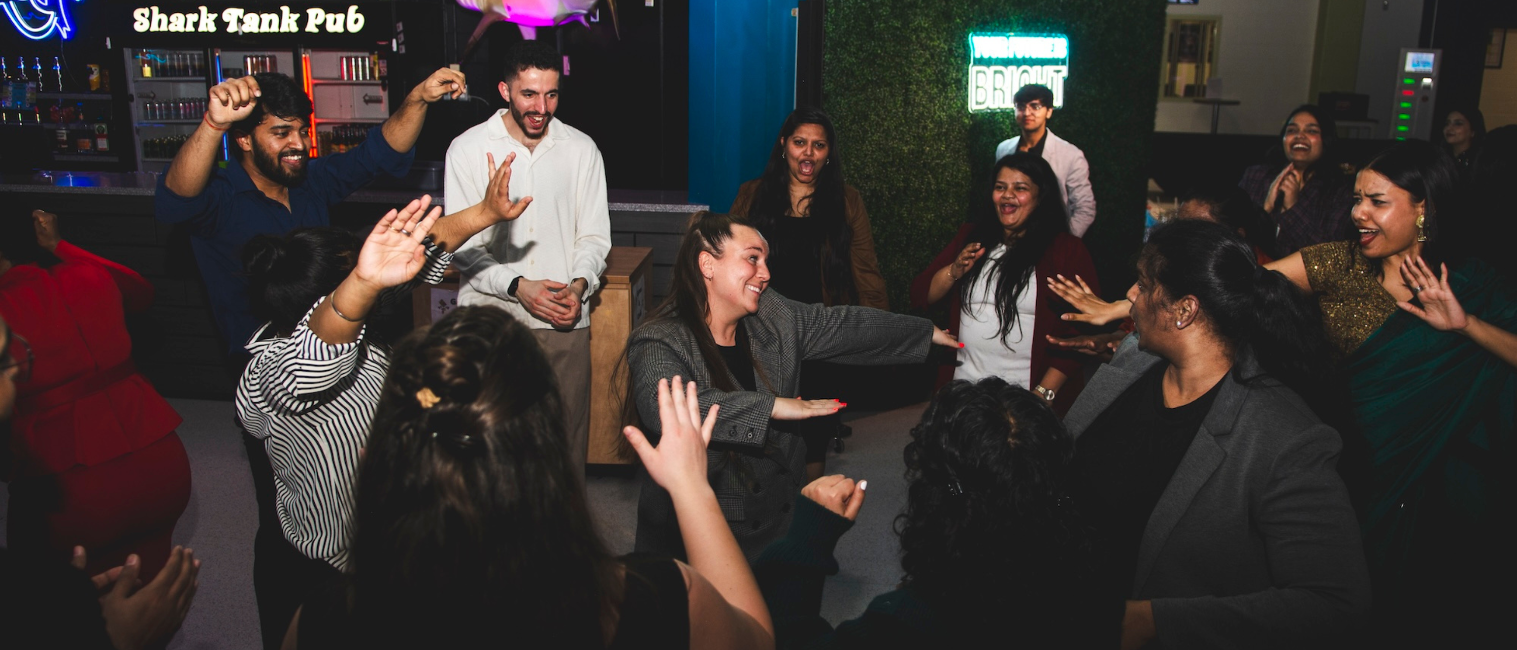 A lively group of people dancing and laughing inside a pub. A neon sign reading "Shark Tank Pub" glows in the background.