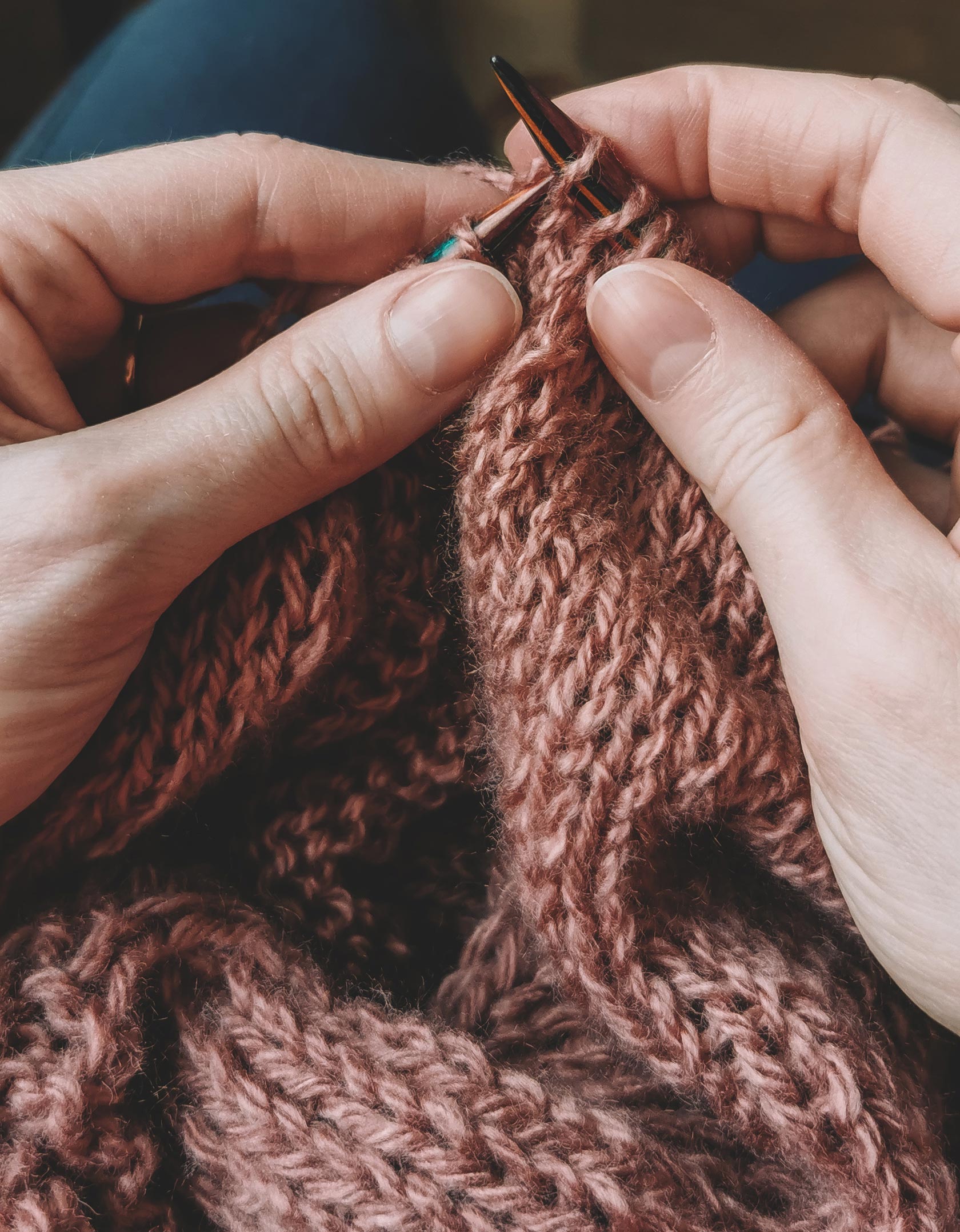 A close-up of hands knitting with brown wool yarn and knitting needles, creating a textured fabric.