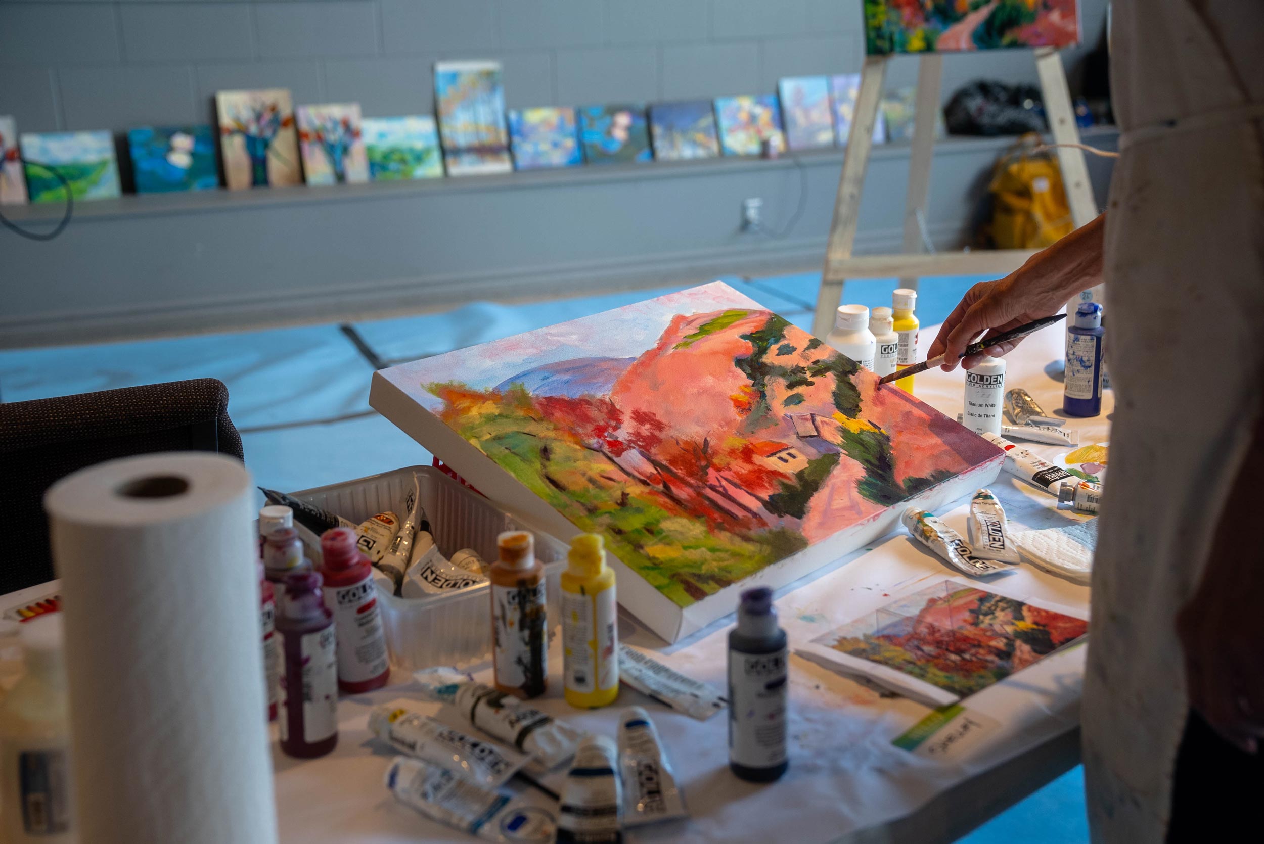 A close-up of a painting in progress on an easel, with an artist’s hand holding a paintbrush touching the canvas. The painting features vibrant red and green colors, possibly depicting a landscape or abstract design. Various art supplies, including tubes of paint, a palette knife, and paper towels, are around the canvas. In the background, additional paintings are propped up for display.