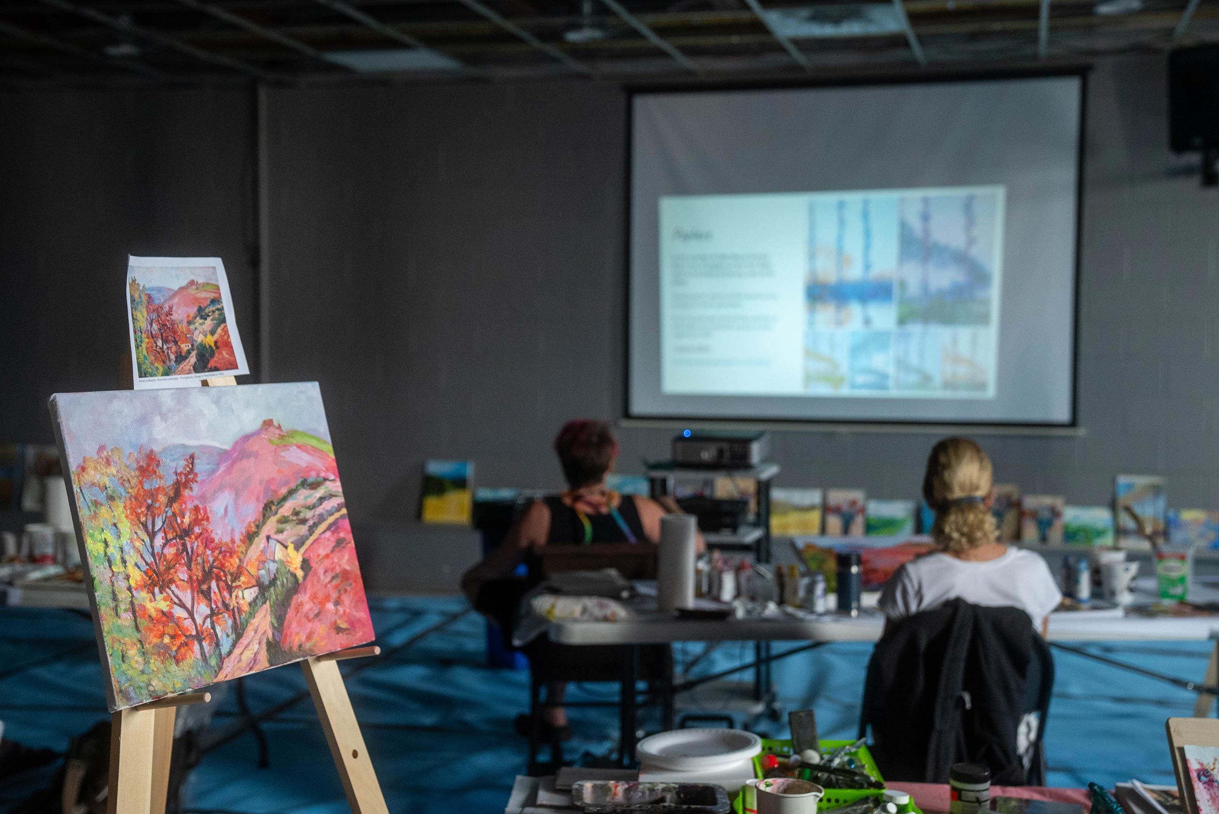 An art class setting with individuals seated at tables, each with a canvas and painting supplies. In the foreground, a canvas on an easel displays a colourful landscape with red foliage. In the background, a projector screen shows a presentation slide titled ‘Palette,’ indicating an instructional session.