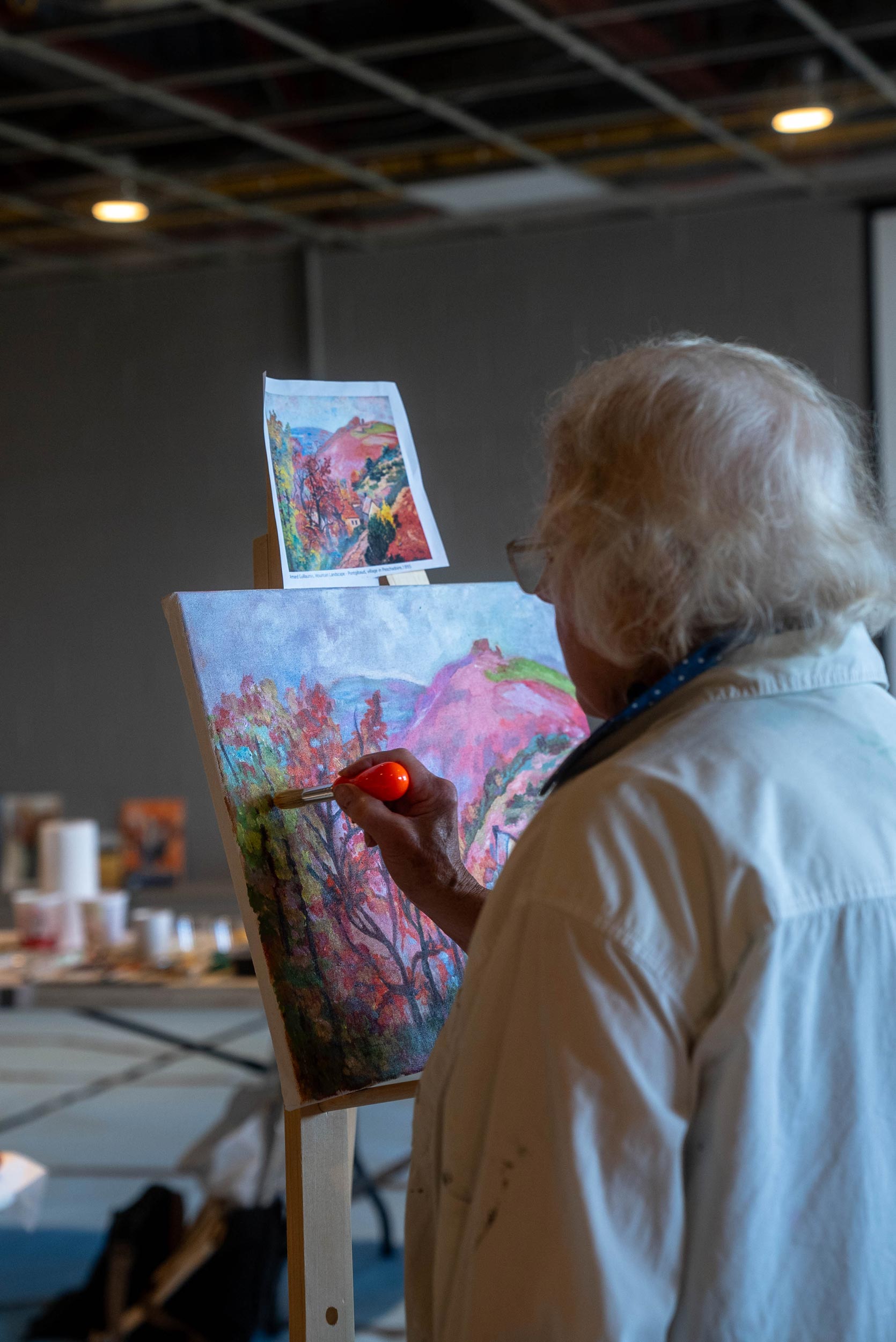 An individual painting on a canvas set on an easel, replicating a smaller picture of a colourful landscape with trees and foliage in red and green hues. The canvas shows progress in capturing the scene from the reference image, with similar colors and composition being applied. The scene is indoors with artificial lighting.