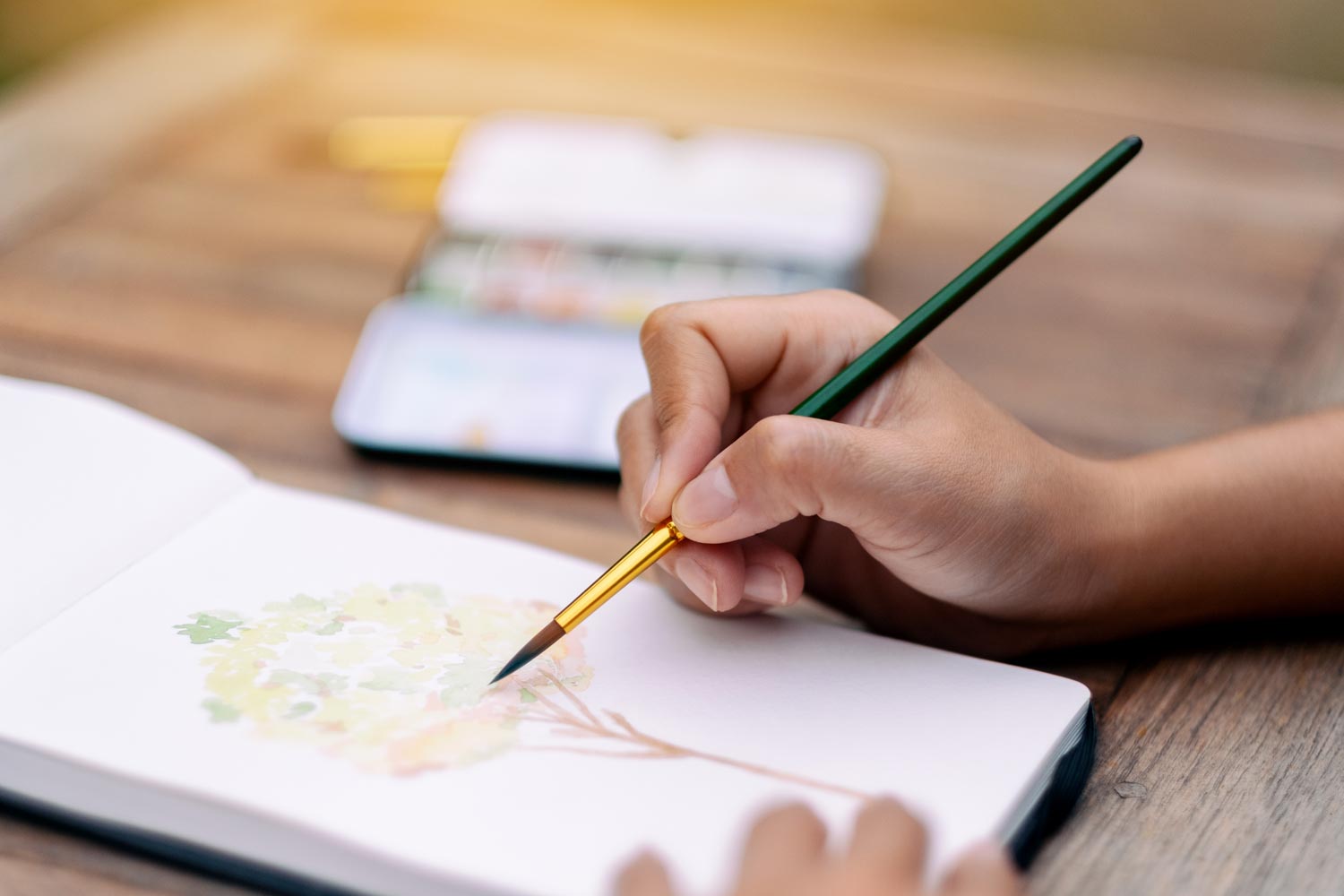 A close-up of a person’s hand holding a paintbrush, painting green foliage with yellow accents on white paper. In the background, there is a blurred notebook and a yellow pen or pencil.