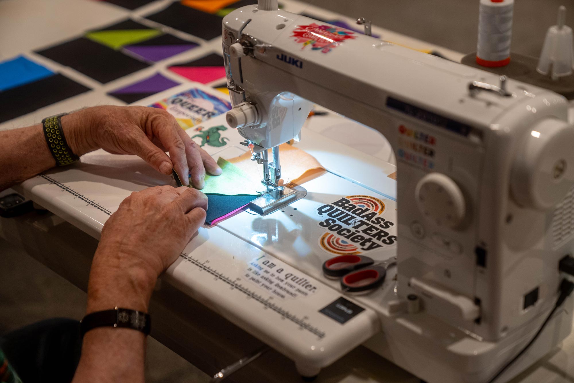 A close-up of a person’s hands using a white sewing machine to stitch colourful fabric patches, likely part of a quilting project. The machine has several stickers applied to it.