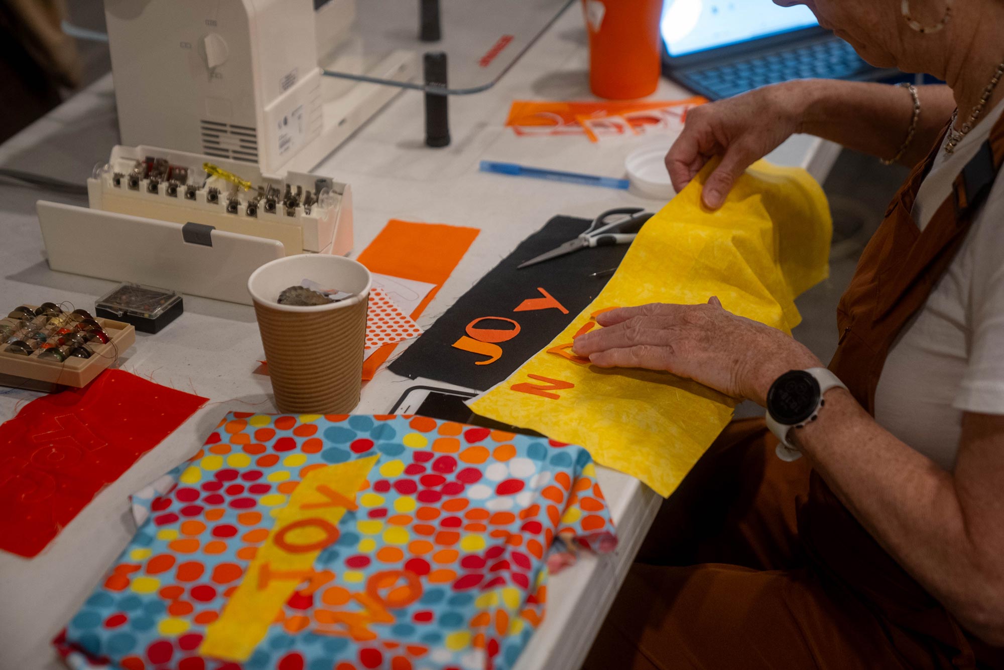 A person’s hands work on a sewing project with bright yellow fabric and red letters spelling ‘JOY’ on a black fabric background. To the left, there are more colorful fabrics, a sewing machine with spools of thread, and a paper cup filled with pins or needles.