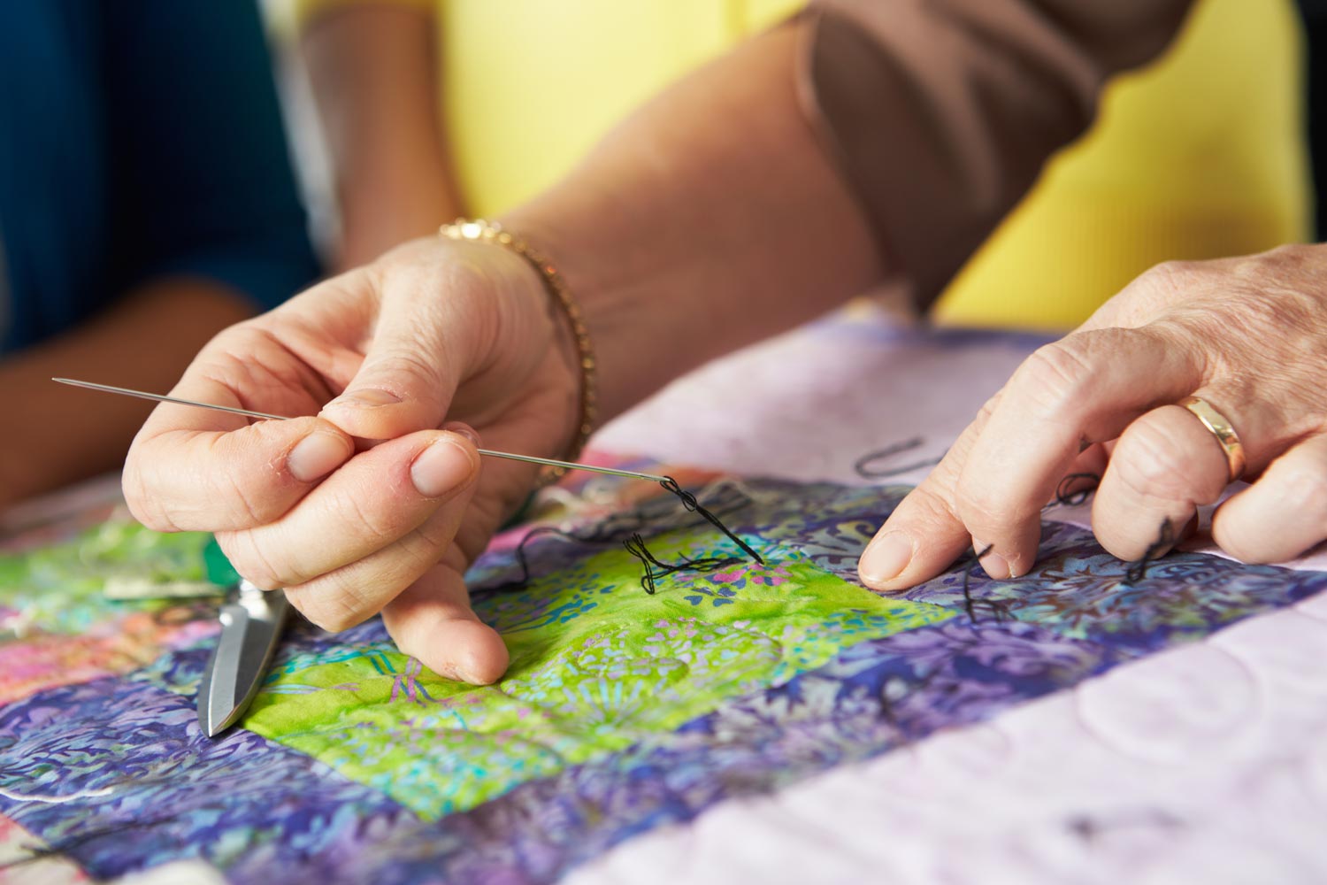 A close-up of hands working on a quilt, using a needle and thread to stitch through layers of colourful fabric.