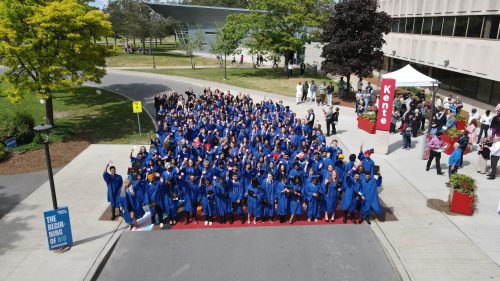 A drone photo above a group of Loyalist graduates in blue convocation gowns out front of Loyalist's Belleville campus Kente building.