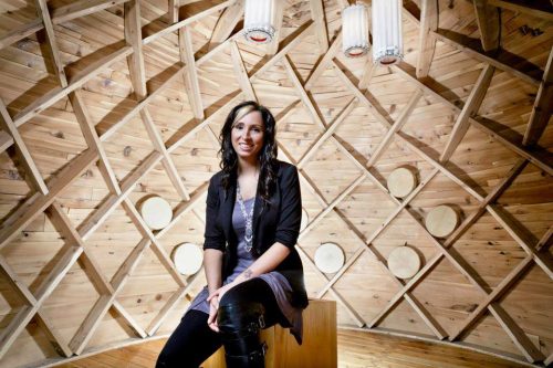 An image of Pam Palmater, sitting in a wooded room, smiling.