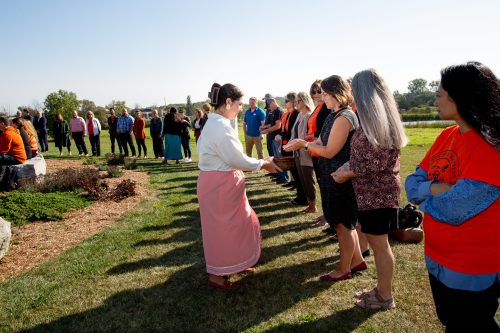 A group of people are gathered together in a semi-circle and a woman is going around with a bowl as part of a Truth and Reconciliation week ceremony. Several people are wearing orange shirts.