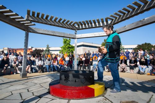 A closeup image of the firepit of the A’nó:wara Learning Circle, with a student holding a bowl standing over the fire.