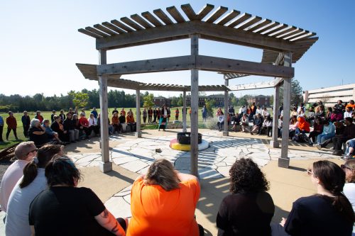 The A’nó:wara Learning Circle at Loyalist College, with people sitting all around the circle and a firepit in the middle.