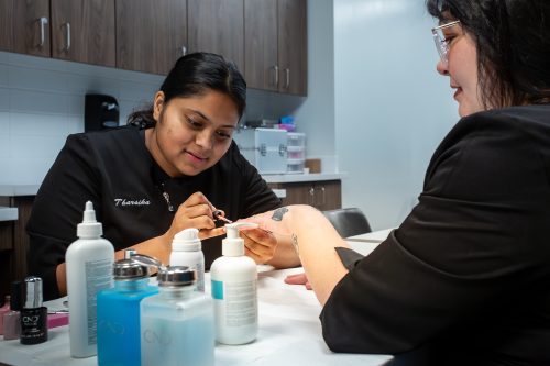 An esthetics and spa management student is painting a mock client's nails. The student and mock client are both wearing black uniforms and smiling.