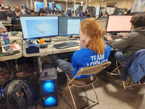 A Loyalist student wearing a blue Team Loyalist shirt, sitting in front of a computer. On the screen are 3D shapes being generated. In the background there are several more rows of people at computers.