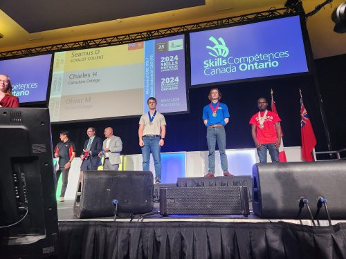 Three students on a stage at a Skills Canada Ontario competition with medals, standing before an audience. A screen in the background shows Seamus D of Loyalist College won the gold medal.