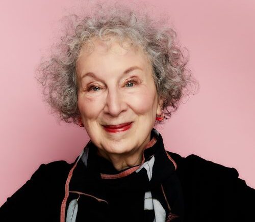 A headshot of Margaret Atwood, smiling with her arms crossed in front of her, in front of a light pink background.