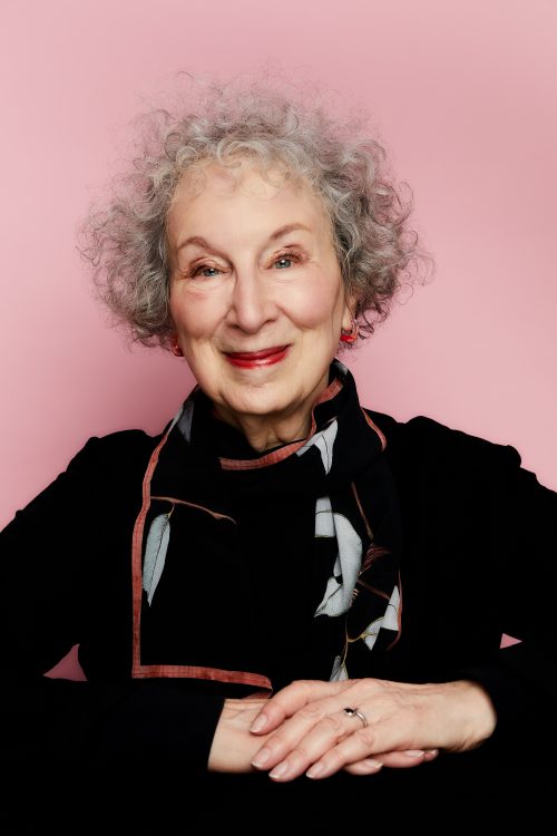 A headshot of Margaret Atwood, smiling with her arms crossed in front of her, in front of a light pink background.