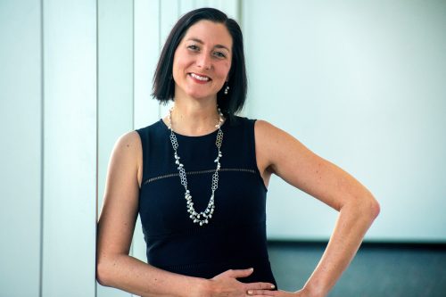 Headshot of Amanda Baskwill, Dean of Health, Human and Justice Studies, wearing a sleeveless black dress and a long silver necklace. She is standing with one arm resting on her waist and leaning slightly to the side, set against a light-coloured, minimalistic background.