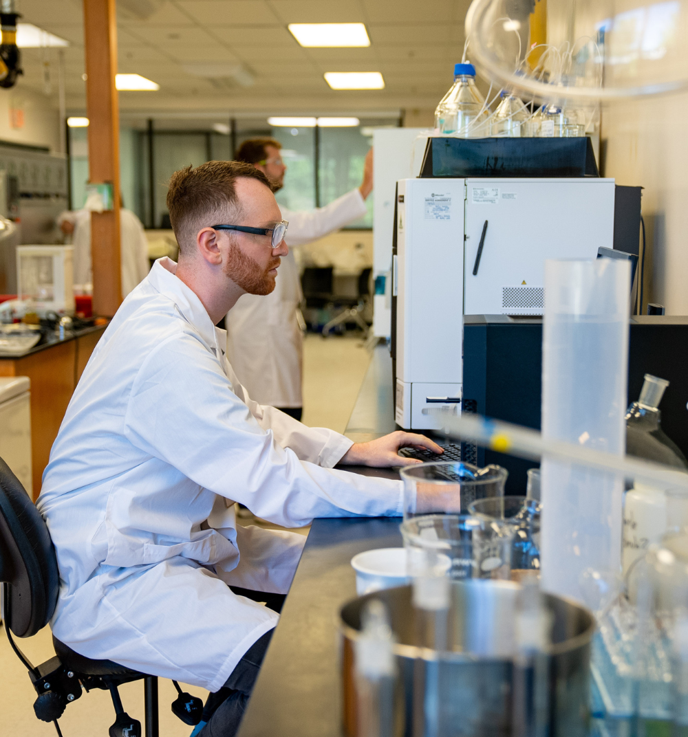 A man in a lab coat is seated at a work station. Around him is the rest of a research lab with beakers and other equipment.