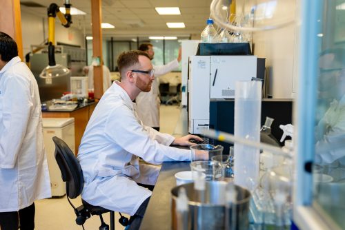 A man in a lab coat is seated at a work station. Around him is the rest of a research lab with beakers and other equipment.