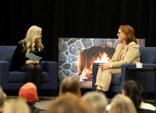 Arlene Dickinson and Jill Raycroft sitting on sofa chairs on a raised platforms in front of an artificial fire.