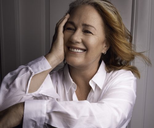 A headshot of Arlene Dickinson, smiling with her hand resting on the side of her face. The background is a white wood panelling.