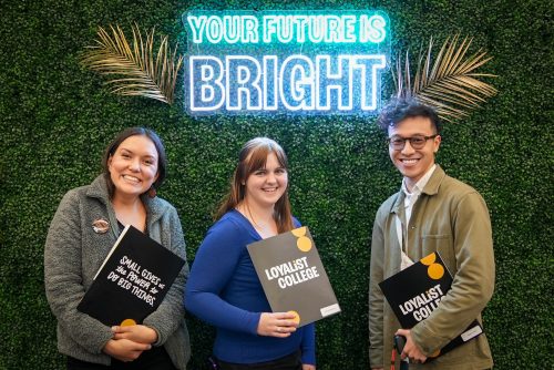 Three students posing with Loyalist Awards folders in front of a neon sign saying "Your Future Is Bright."