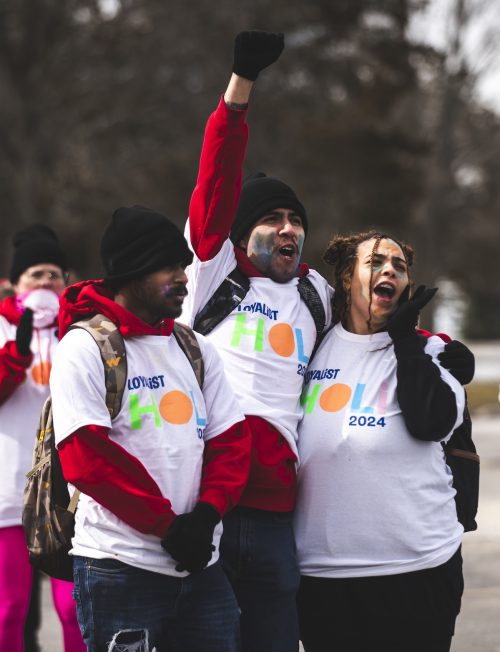 Three Loyalist students cheering and huddled together, all wearing the same Loyalist Holi 2024 white T-shirt over their clothes.