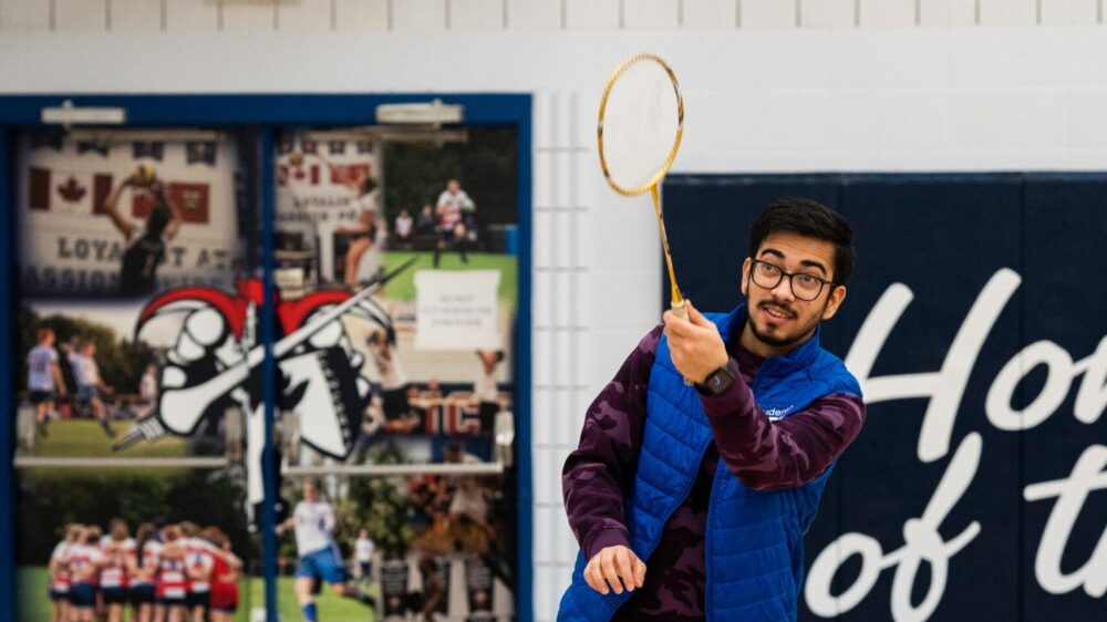 Man playing badminton indoors, he has just hit a birdie with his racket. He is wearing glasses, a blue vest, and dark pants. He is in a gym with polished wooden floors, and behind him is a wall with sports banners and murals depicting various sports.