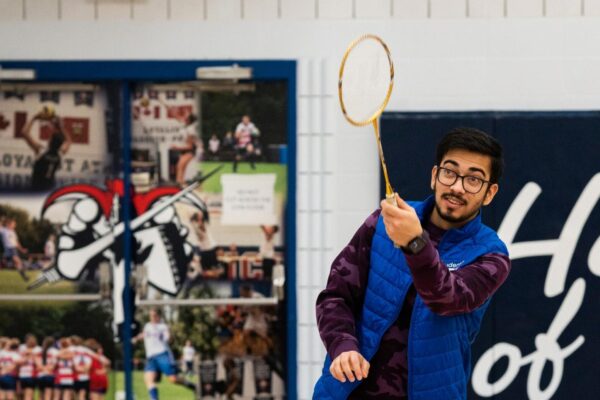 Man playing badminton indoors, he has just hit a birdie with his racket. He is wearing glasses, a blue vest, and dark pants. He is in a gym with polished wooden floors, and behind him is a wall with sports banners and murals depicting various sports.