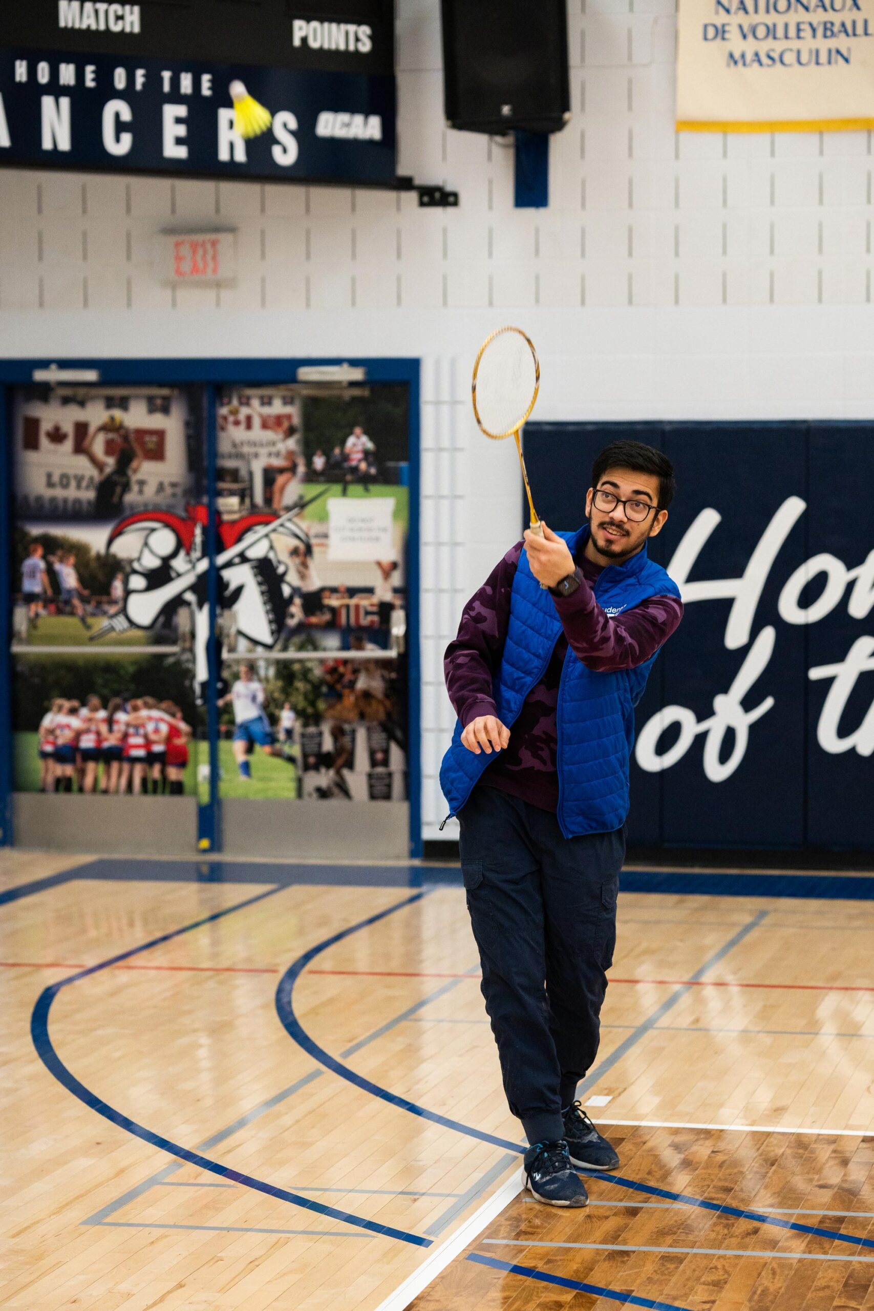 Man playing badminton indoors, he has just hit a birdie with his racket. He is wearing glasses, a blue vest, and dark pants. He is in a gym with polished wooden floors, and behind him is a wall with sports banners and murals depicting various sports.