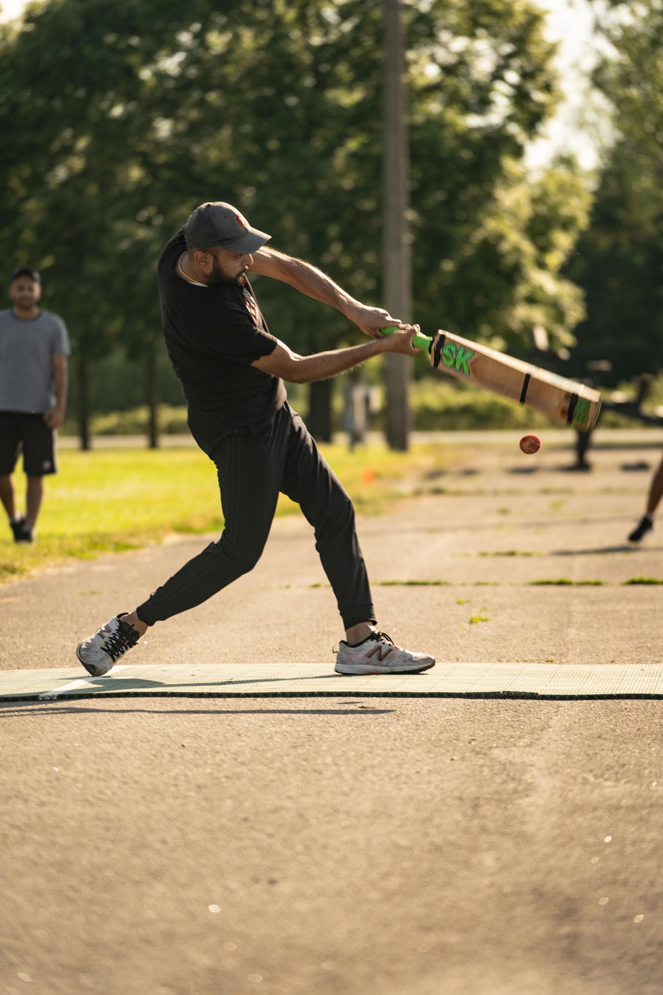 Man swinging a cricket bat during an outdoor game, focused on hitting the ball. He is wearing a black shirt, black pants, and a hat, with another player visible in the background. The game is taking place on a sunny day with trees and grass in the distance.