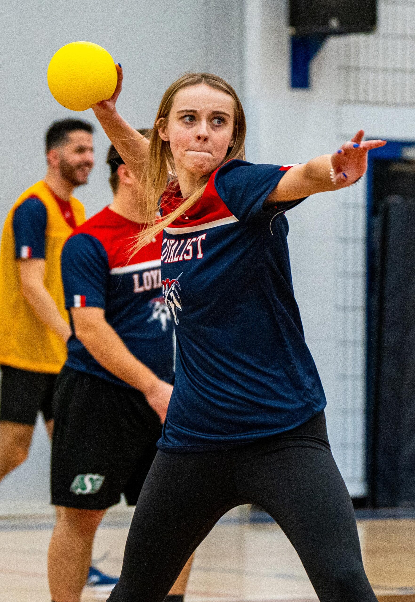 Young woman wearing a Loyalist Lancers t-shirt, winding up to throw a yellow ball during an indoor dodgeball game. She is focused on her throw, with other players in the background, one who is wearing a yellow vest.