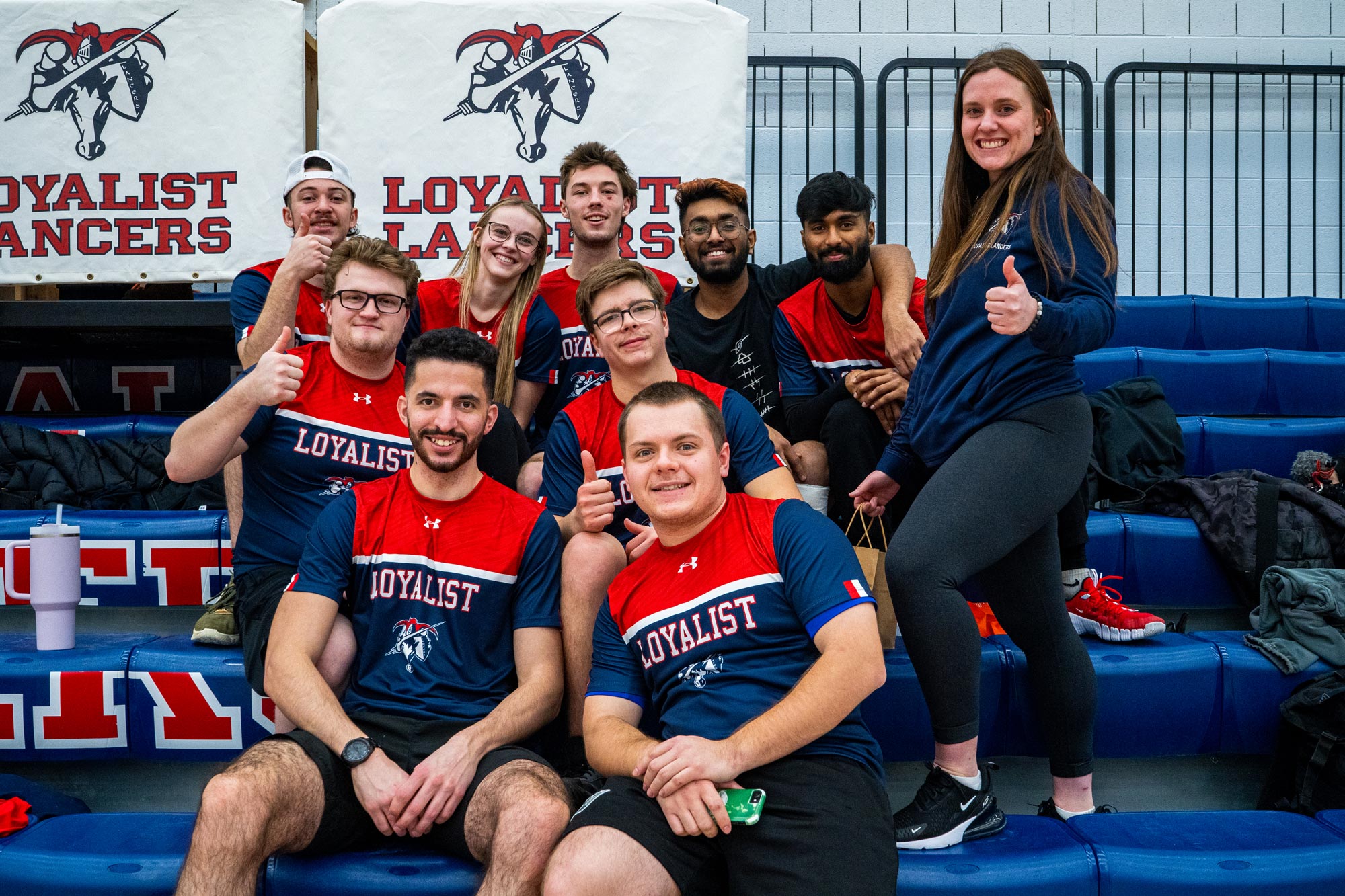 Group of Loyalist students sitting on bleachers and smiling for the camera. They are wearing red and blue Loyalist Lancers jerseys, with one woman standing on the right giving a thumbs-up. Behind them, a banner with the Loyalist Lancers logo is displayed. The group appears relaxed and cheerful in a gymnasium setting.