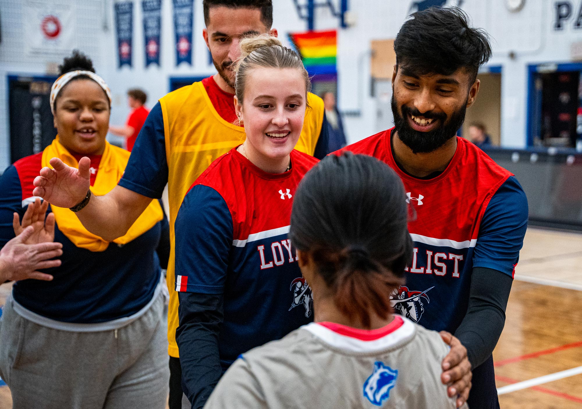 Group of students wearing Loyalist Lancers-branded sports jerseys, smiling at each other. A woman in a red and blue jersey is at the centre, shaking hands with another player. Other teammates are in the background, including one wearing a yellow vest. The scene takes place indoors in a gymnasium with sports banners and a rainbow flag visible in the background.