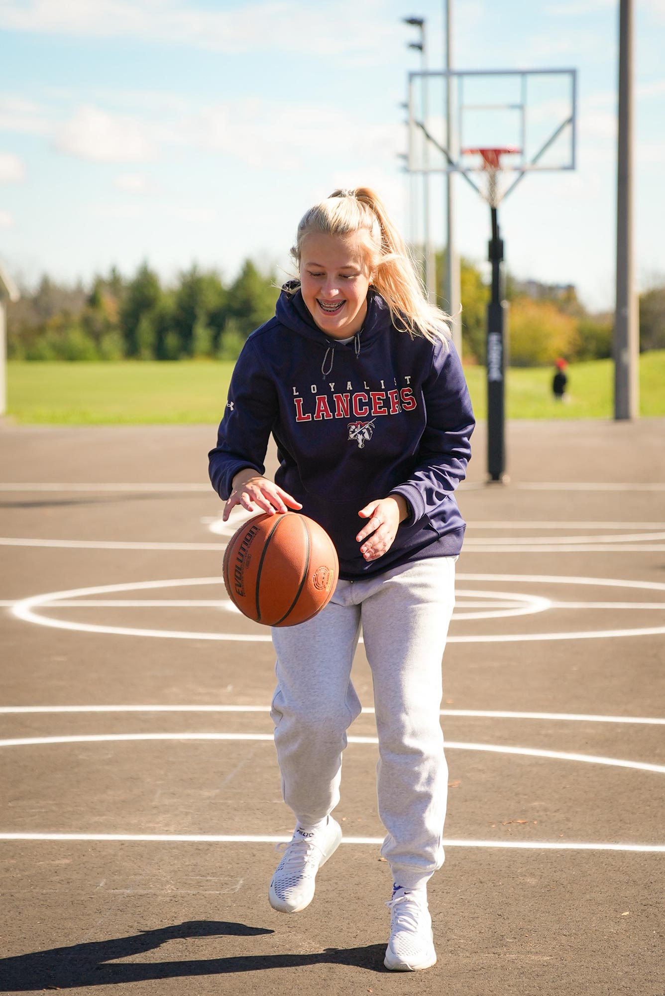 Young woman dribbling a basketball outdoors on a sunny day. She is wearing a navy 'Loyalist Lancers' hoodie and grey sweatpants, smiling as she plays on a basketball court. A basketball hoop is visible in the background, with trees and a green field in the distance.