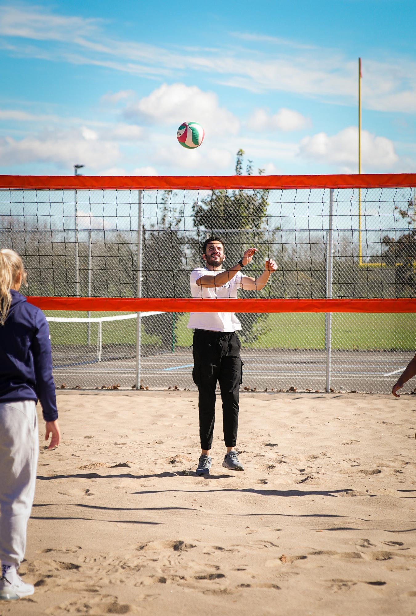 Outdoor volleyball game on a sand court with a clear blue sky. A man in a white shirt is standing on the other side of the volleyball net, preparing to hit the ball, which is in the air above him. Another person, seen from behind, is on the left side of the image. A chain-link fence and goalpost are visible in the background.