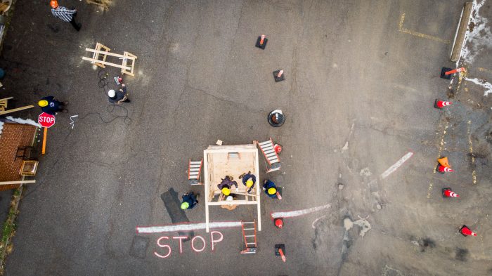 An aerial view of construction students working on a small wooden structure in a parking lot. Several people in hard hats are gathered around the framework, with tools and ladders nearby. The word 'STOP' is painted on the ground, along with traffic cones and a stop sign visible in the scene.