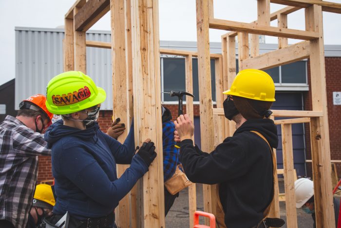 A group of construction students working on a wooden framework outdoors. They are wearing hard hats and face masks, with one person using a hammer while another holds the structure in place. The scene takes place near a building with metal siding and windows in the background.