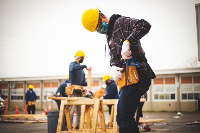 Construction student wearing a yellow hard hat, mask, and tool belt, adjusting tools while working outdoors. In the background, other students, also in hard hats and masks, are working on a project at a wooden sawhorse. The scene takes place in a parking lot near a building.